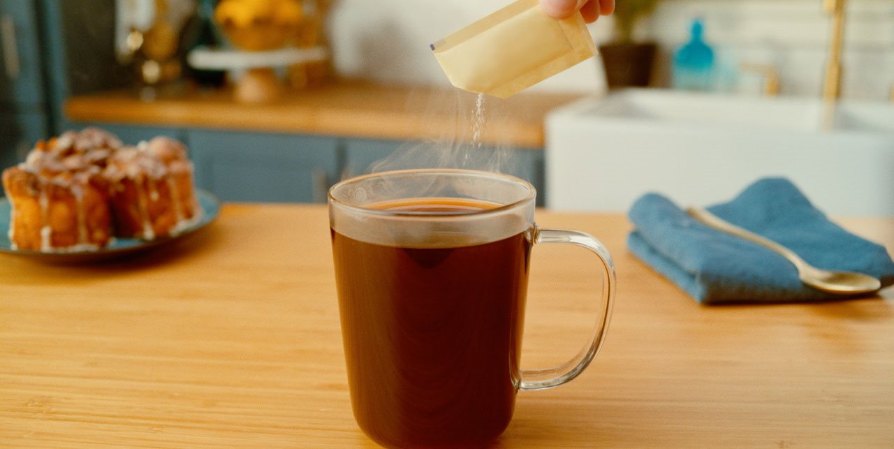 Hand pouring Splenda packet into coffee on a wooden kitchen counter