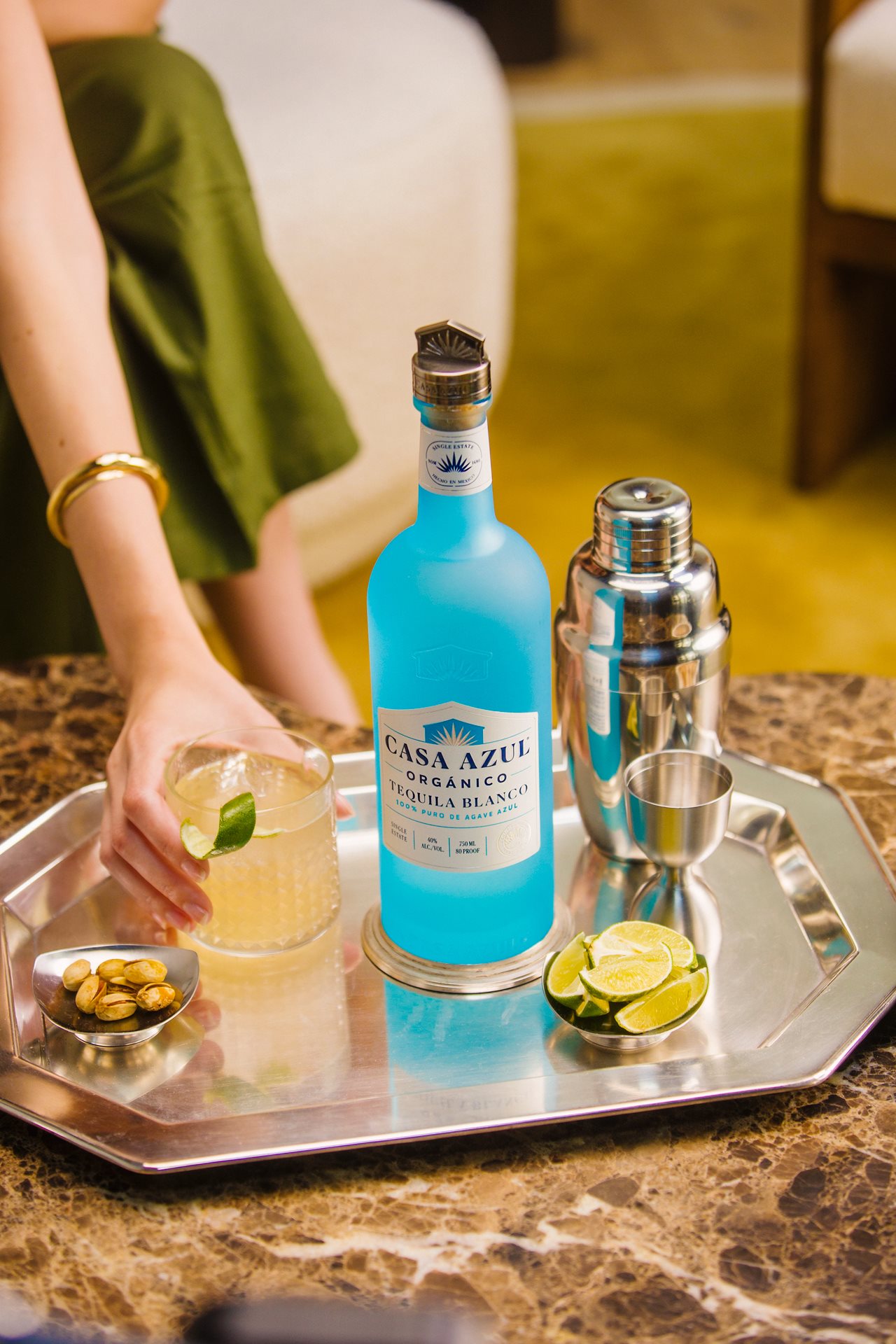 Close up of a woman holding a drink glass next to a bottle of Casa Azul Blonco